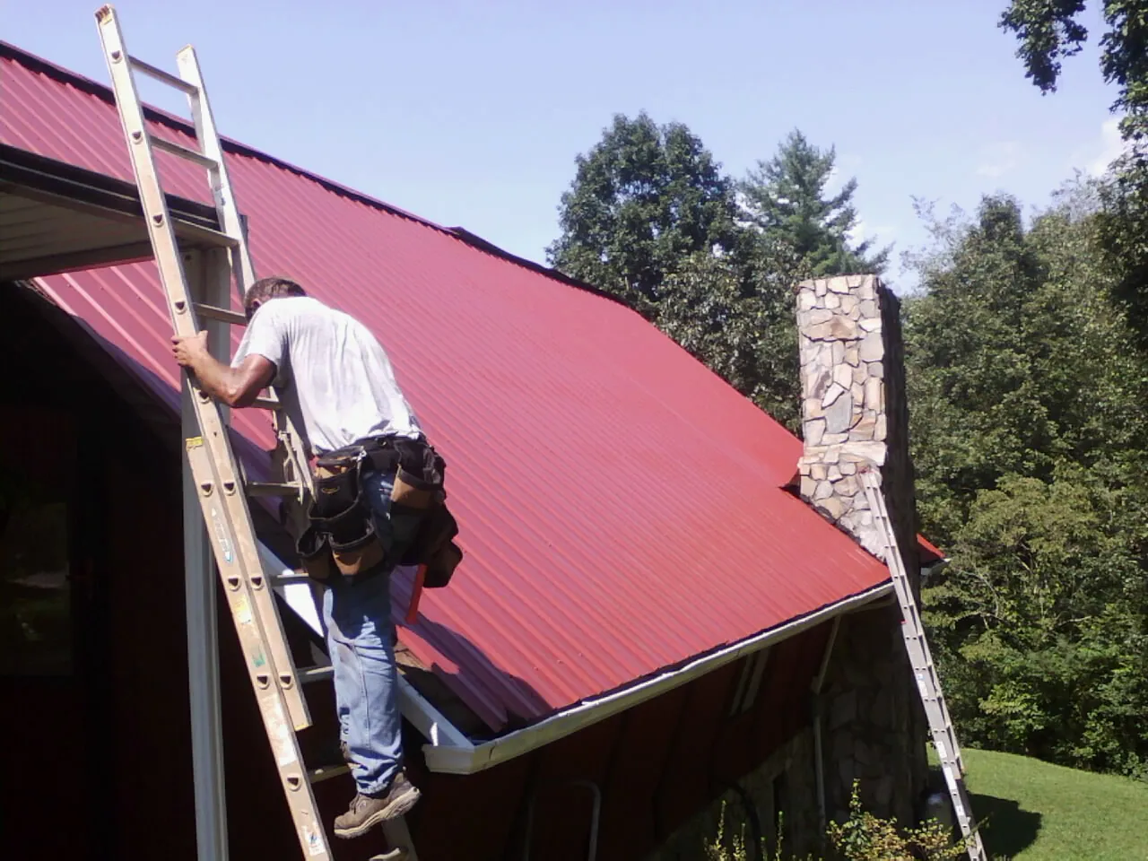 Roofer on a ladder beside a red metal roof and stone chimney — Soffit & Fascia Repair by Roofing Brothers in Moores Hill
